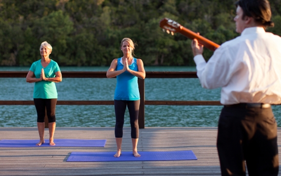 Two women do yoga on a dock overlooking the water while a man plays guitar near them.