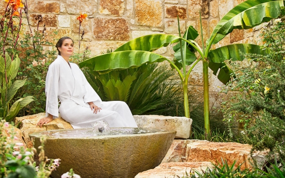 A woman wearing a robe sits in a garden near a small bubbling fountain.